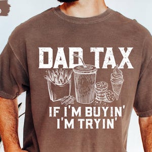 May include: Espresso-colored t-shirt with the text "DAD TAX" above illustrations of fries, a drink, cookies, and ice cream. The phrase "IF I'M BUYIN' I'M TRYIN'" is below the illustrations. The shirt is a casual wear item.