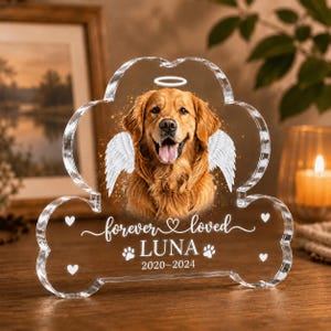 May include: A clear acrylic paw-shaped memorial plaque featuring a golden retriever portrait with white angel wings and a halo. The text reads forever loved, LUNA, and 2020-2024, accented with small white heart and paw print icons.