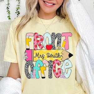 May include: A light yellow t-shirt with the words "FRONT OFFICE Mrs. Smith" in colorful, playful lettering. The design includes hearts, pencils, and flowers. The shirt is a comfortable, casual style.