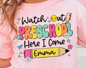 May include: A light pink t-shirt featuring a colorful graphic with the text "Watch out PRESCHOOL Here I Come" and the name "Emma" written on a yellow pencil illustration. The design includes a smiley face, lightning bolt, and stars.