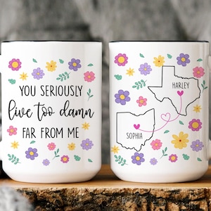 May include: Two white ceramic mugs with black handles and rims. One mug has the text "YOU SERIOUSLY live too damn FAR FROM ME" with floral accents. The other mug features state outlines with names and floral designs.