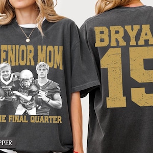 May include: A charcoal gray t-shirt featuring a vintage-style graphic on the front with the text SENIOR MOM and THE FINAL QUARTER, alongside a black and white photo of three football players. The back displays the name BRYAN and the number 15.