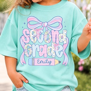 May include: A light turquoise t-shirt featuring a graphic with a large pink and blue bow above the text Second Grade in a mix of gingham and floral patterns. Below the text is a pencil graphic with the name Emily printed in blue script.