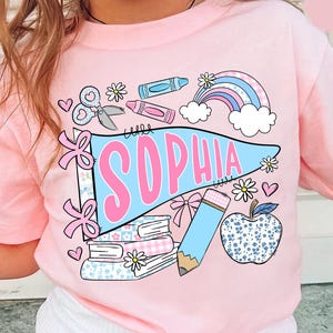 May include: A light pink t-shirt featuring a graphic print of a blue pennant with the name SOPHIA in pink letters. The design includes school-themed illustrations such as a stack of books, a pencil, scissors, a rainbow, and an apple.
