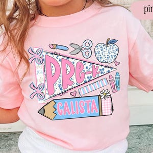 May include: A light pink t-shirt featuring a graphic print of a pennant flag with the text PRE-K, a large pencil with the name CALISTA, a blue floral apple, scissors, a paintbrush, a ruler, and a crayon. The design includes pink and blue accents.