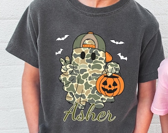 Puede incluir: Camiseta gris oscuro con un fantasma de dibujos animados con un patrón de camuflaje, que lleva una gorra verde y naranja, sosteniendo una linterna de calabaza naranja y haciendo el signo de la paz. El nombre "Asher" está escrito en cursiva. Murciélagos blancos en el fondo.