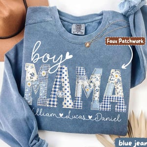 May include: Blue jean-colored sweatshirt with the words "boy MAMA" in white script and patchwork letters. The patchwork letters feature floral, polka dot, and gingham patterns. The names William, Lucas, and Daniel are printed below.