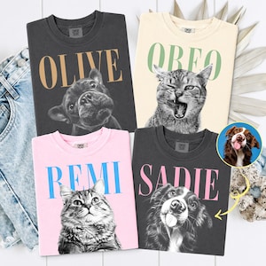 May include: Four crewneck t-shirts featuring custom pet portraits and names. One charcoal shirt says OLIVE with a French Bulldog, one cream shirt says OREO with a cat, one pink shirt says REMI with a cat, and one charcoal shirt says SADIE with a dog.