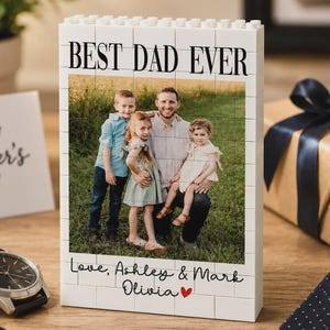 May include: A white rectangular block display constructed from interlocking plastic bricks featuring a printed photograph of a father and three children in a grassy field. The text BEST DAD EVER appears at the top, with Love, Ashley & Mark Olivia and a red heart at the bottom.