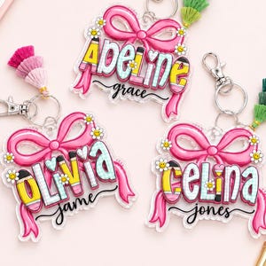 May include: Three personalized acrylic keychains featuring a large pink bow design with small yellow daisy accents. Each keychain displays a name in colorful, pencil-themed lettering with a tassel attached to a silver metal lobster clasp hardware.