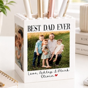 May include: A white block-style pen holder with a family photo and the text "BEST DAD EVER." The holder contains four pens in gold, rose gold, and white. The bottom of the holder has the text "Love, Ashley & Mark Olivia ❤️."