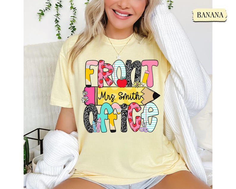 May include: A light yellow t-shirt with the words "FRONT OFFICE Mrs. Smith" in colorful, playful lettering. The design includes hearts, pencils, and flowers. The shirt is a comfortable, casual style.