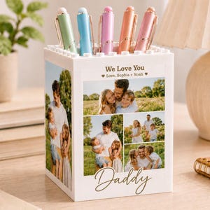 May include: A white pen holder with a personalized photo collage and the text "We Love You" and "Daddy". The pen holder contains five pens in pastel colors: green, blue, pink, orange, and pink. A thoughtful gift.