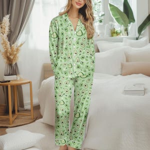 Könnte beinhalten: Hellgrünes Pyjama-Set mit Avocadomuster. Das langärmlige Oberteil und die Hose zeigen ein sich wiederholendes Design aus Avocados, Blättern und kleinen weißen Blumen. Weiße Paspeln säumen die Ränder des Pyjamas.