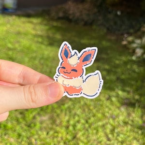 Op de afbeelding: Een sticker met een cartoon Flareon-personage. De sticker heeft een witte rand en toont het personage in tinten oranje, cr&egrave;me en blauw. Het personage lacht met zijn staart omhoog. De sticker wordt in een hand gehouden tegen een groene achtergrond.