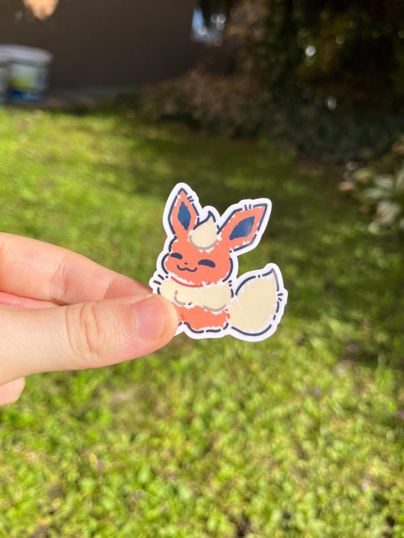 Op de afbeelding: Een sticker met een cartoon Flareon-personage. De sticker heeft een witte rand en toont het personage in tinten oranje, cr&egrave;me en blauw. Het personage lacht met zijn staart omhoog. De sticker wordt in een hand gehouden tegen een groene achtergrond.