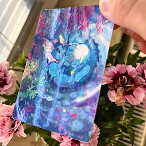 May include: A holographic card featuring a blue cartoon creature in a waterfall setting. The card has a glossy finish with iridescent accents. Pink carnations and green foliage are in the foreground.