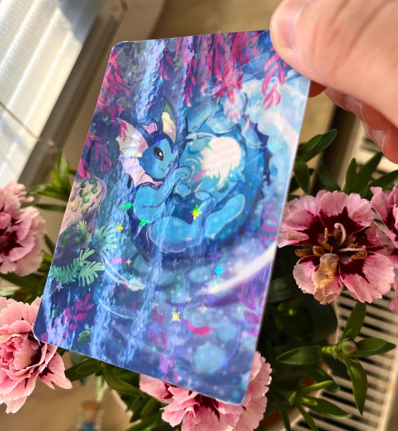 May include: A holographic card featuring a blue cartoon creature in a waterfall setting. The card has a glossy finish with iridescent accents. Pink carnations and green foliage are in the foreground.