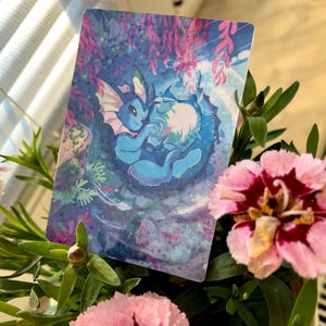 May include: A rectangular card featuring a blue cartoon creature with pink accents, set against a blue and purple underwater scene. The card is surrounded by pink and red flowers and green foliage.