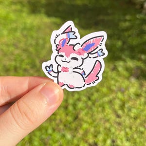 Op de afbeelding: Een gestanste sticker met een cartoon Sylveon-personage. De sticker heeft een witte achtergrond met een roze, blauw en zwart ontwerp. Het personage lacht en zwaait. De sticker wordt door een hand tegen een groene achtergrond gehouden.