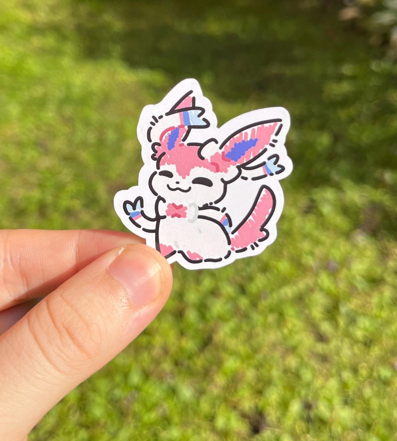Op de afbeelding: Een gestanste sticker met een cartoon Sylveon-personage. De sticker heeft een witte achtergrond met een roze, blauw en zwart ontwerp. Het personage lacht en zwaait. De sticker wordt door een hand tegen een groene achtergrond gehouden.