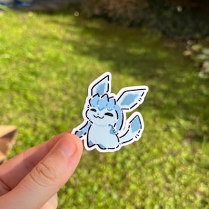 Op de afbeelding: Een lichtblauwe sticker met een cartoonfiguur met grote oren en een pluizige staart. De sticker heeft een witte omtrek en wordt door een hand vastgehouden tegen een groene, grasrijke achtergrond.