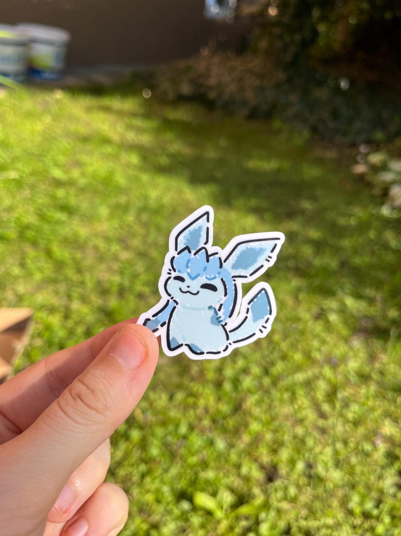Op de afbeelding: Een lichtblauwe sticker met een cartoonfiguur met grote oren en een pluizige staart. De sticker heeft een witte omtrek en wordt door een hand vastgehouden tegen een groene, grasrijke achtergrond.