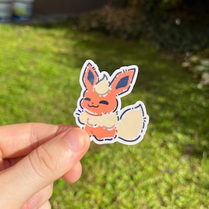Op de afbeelding: Een sticker met een cartoon Flareon-personage. De sticker heeft een witte rand en toont het personage in tinten oranje, cr&egrave;me en blauw. Het personage lacht met zijn staart omhoog. De sticker wordt in een hand gehouden tegen een groene achtergrond.