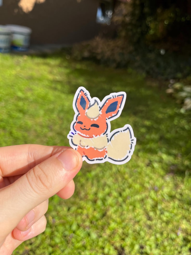Op de afbeelding: Een sticker met een cartoon Flareon-personage. De sticker heeft een witte rand en toont het personage in tinten oranje, cr&egrave;me en blauw. Het personage lacht met zijn staart omhoog. De sticker wordt in een hand gehouden tegen een groene achtergrond.