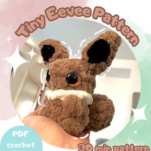 Puede incluir: Un peluche Eevee de ganchillo marrón con puntas de orejas negras y ojos grandes y brillantes de color naranja. El peluche tiene un cuello blanco y se sostiene en una mano. El texto en la imagen dice "Tiny Eevee Pattern", "PDF crochet pattern", y "30 min pattern Bestseller!"