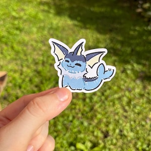 Op de afbeelding: Een sticker met een cartoon Vaporeon-personage in tinten blauw, wit en zwart. De sticker heeft een witte rand en wordt tegen een vage groene achtergrond gehouden.
