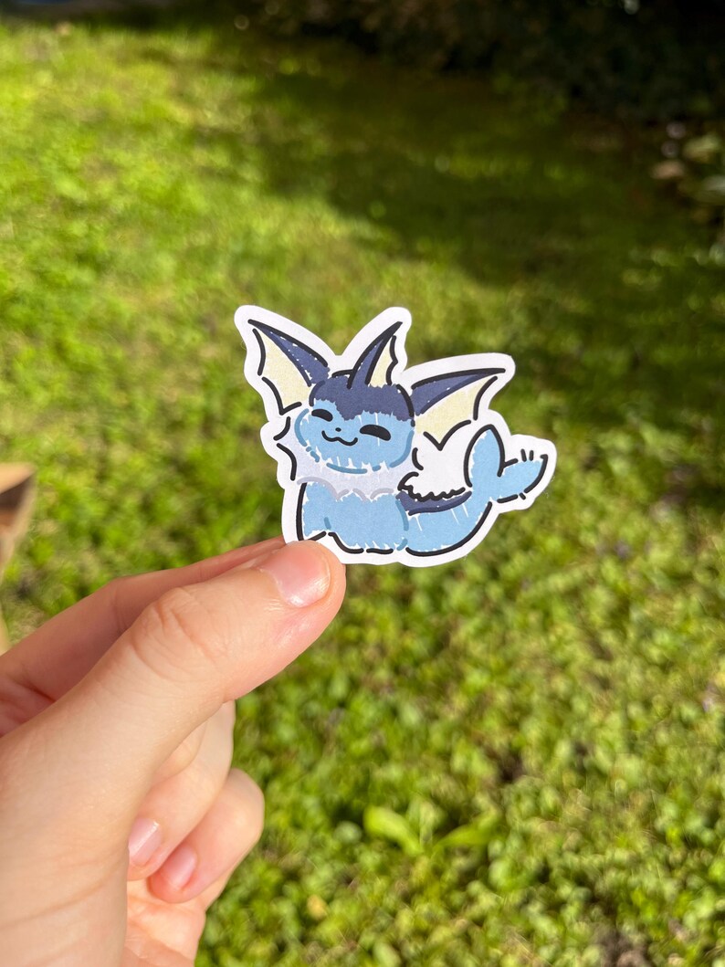 Op de afbeelding: Een sticker met een cartoon Vaporeon-personage in tinten blauw, wit en zwart. De sticker heeft een witte rand en wordt tegen een vage groene achtergrond gehouden.