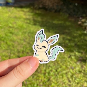 Op de afbeelding: Een sticker met een stripfiguur met een cr&egrave;mekleurig lichaam, groene accenten en een lachend gezicht. Het personage heeft bladvormige kenmerken en is zwart omlijnd. De sticker wordt tegen een achtergrond van groen gras gehouden.