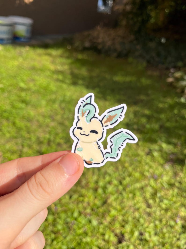 Op de afbeelding: Een sticker met een stripfiguur met een cr&egrave;mekleurig lichaam, groene accenten en een lachend gezicht. Het personage heeft bladvormige kenmerken en is zwart omlijnd. De sticker wordt tegen een achtergrond van groen gras gehouden.