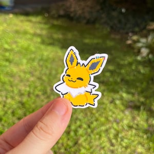 Op de afbeelding: Een gele en witte sticker met een stripfiguur met grote oren en een donzige kraag. De sticker heeft een zwarte omtrek en wordt tegen een groene, grasrijke achtergrond gehouden.