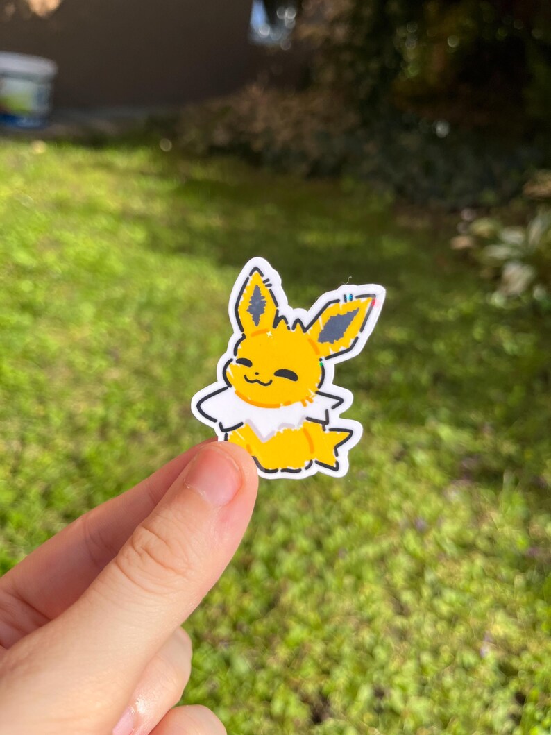 Op de afbeelding: Een gele en witte sticker met een stripfiguur met grote oren en een donzige kraag. De sticker heeft een zwarte omtrek en wordt tegen een groene, grasrijke achtergrond gehouden.
