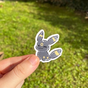 Op de afbeelding: Een sticker met het Umbreon-personage, een zwart Pok&eacute;mon met gele markeringen. De sticker toont het personage glimlachend, met gele accenten op zijn oren en staart. Het wordt tegen een achtergrond van groen gras gehouden.