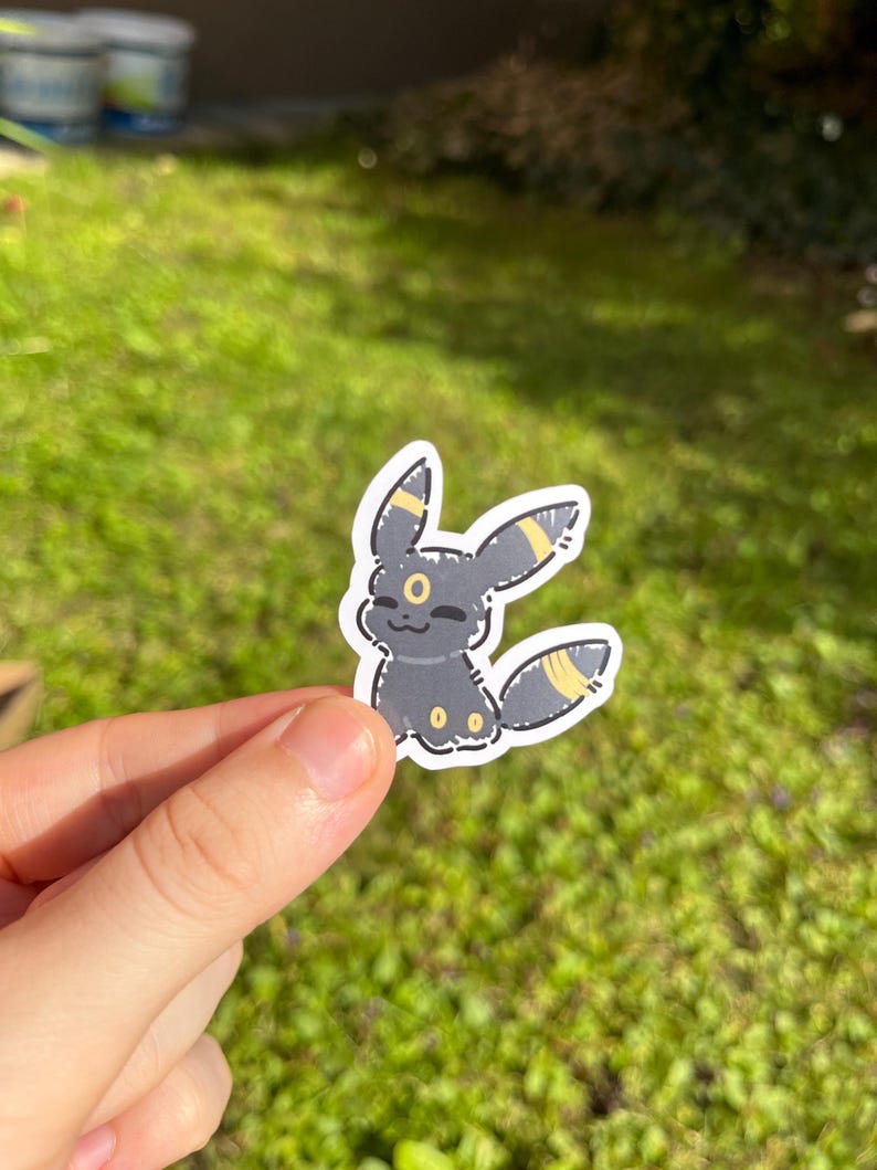 Op de afbeelding: Een sticker met het Umbreon-personage, een zwart Pok&eacute;mon met gele markeringen. De sticker toont het personage glimlachend, met gele accenten op zijn oren en staart. Het wordt tegen een achtergrond van groen gras gehouden.
