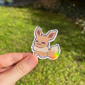 Op de afbeelding: Een sticker met een cartoon Eevee-personage met bruine vacht, een pluizige staart met regenboogaccenten en een glimlachende uitdrukking. De sticker heeft een witte rand en wordt tegen een groene achtergrond gehouden.