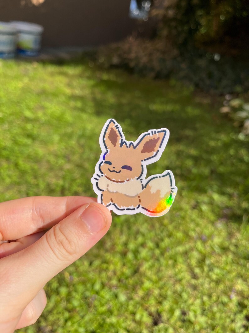 Op de afbeelding: Een sticker met een cartoon Eevee-personage met bruine vacht, een pluizige staart met regenboogaccenten en een glimlachende uitdrukking. De sticker heeft een witte rand en wordt tegen een groene achtergrond gehouden.