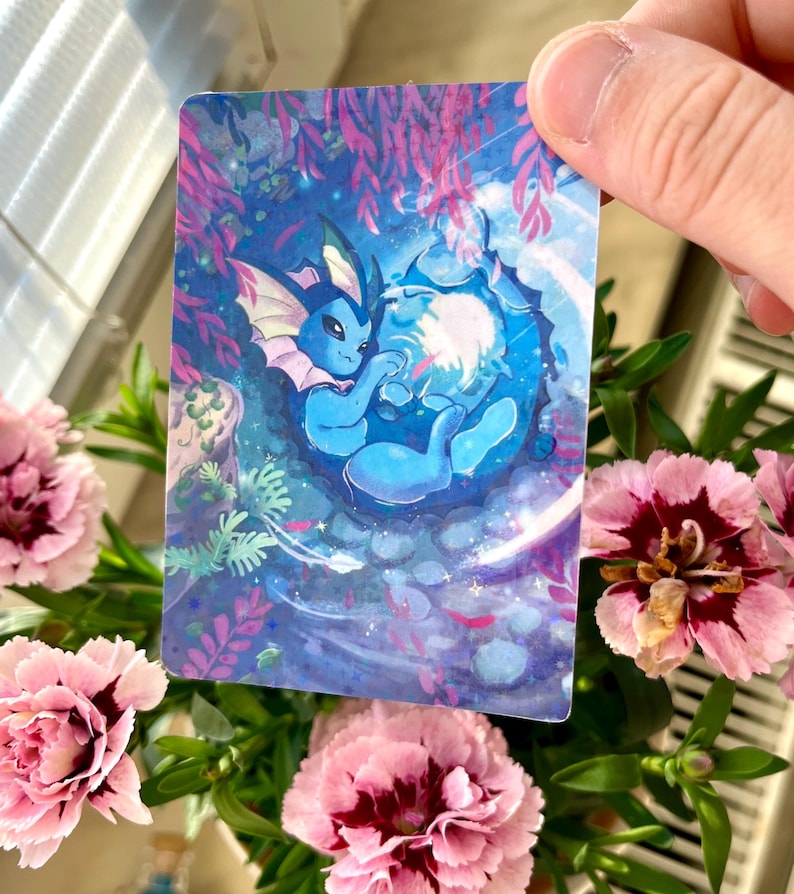 May include: A rectangular card featuring a blue cartoon creature with a light blue and pink design, set against a blue and purple aquatic background. The card is held by a hand, surrounded by pink flowers and green foliage.