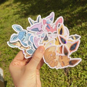 May include: A selection of vibrant stickers depicting cartoon characters. The stickers display diverse designs, including characters with large ears and expressive eyes. The stickers are held in a hand against a backdrop of green grass.