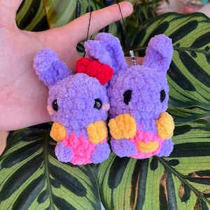Puede incluir: Dos pequeños conejos de peluche tejidos en color violeta con ojos de plástico negro. El conejo izquierdo tiene un lazo rojo brillante y vientre rosa, mientras que el derecho tiene patas y vientre amarillos. Ambos incluyen cordones negros.