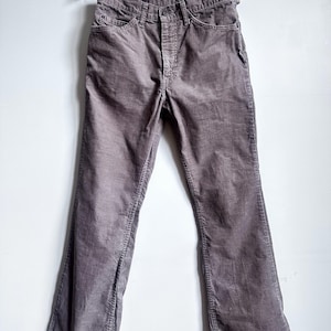 Puede incluir: Un par de pantalones de pana marrones colgados de una percha de madera. Los pantalones tienen un diseño clásico de cinco bolsillos y una pernera acampanada. La tela de pana tiene un aspecto suave y texturizado.