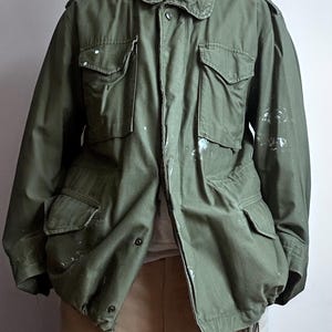 May include: An olive green military jacket with multiple pockets and a button-up front. The jacket features a collar and shoulder epaulettes. There are white paint splatters on the jacket. The jacket is open, showing a light-coloured shirt beneath.