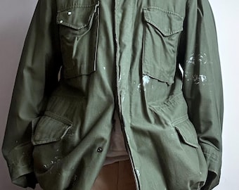 May include: Olive green military-style jacket with multiple front pockets and a button-up closure. The jacket has a collar and shoulder epaulets. White paint splatters are visible on the jacket. The jacket is open, revealing a light-colored shirt underneath.