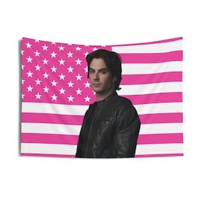 May include: A wall tapestry featuring a portrait of a person in front of a pink and white American flag design. The person is wearing a black leather jacket. The flag has pink stripes and white stars.