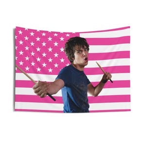 May include: A pink and white American flag tapestry with a person holding drumsticks. The person is wearing a blue t-shirt and appears to be drumming. The flag has white stars on a pink background and pink and white stripes.