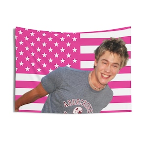 Puede incluir: Un tapiz con diseño de bandera americana rosa y blanca, con una persona sonriente que lleva una camiseta gris de Abercrombie. El tapiz presenta un fondo rosa con estrellas y rayas blancas.