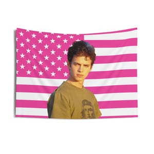 Könnte beinhalten: Ein Wandteppich mit einem rosa und weißen amerikanischen Flaggenmuster. Eine Person mit einem olivgrünen T-Shirt mit Grafik steht vor der Flagge. Der Wandteppich ist rechteckig und aus Stoff.